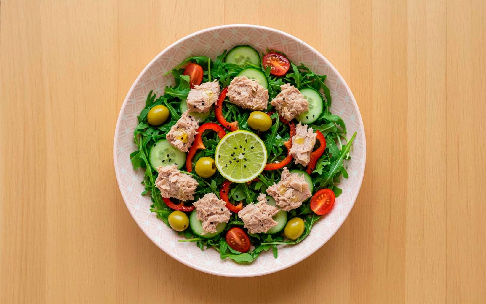 Bowl con ensalada de rúcula, atún, aceitunas verdes, tomate cherry, pepino y pimiento rojo, con rodaja de limón al centro.