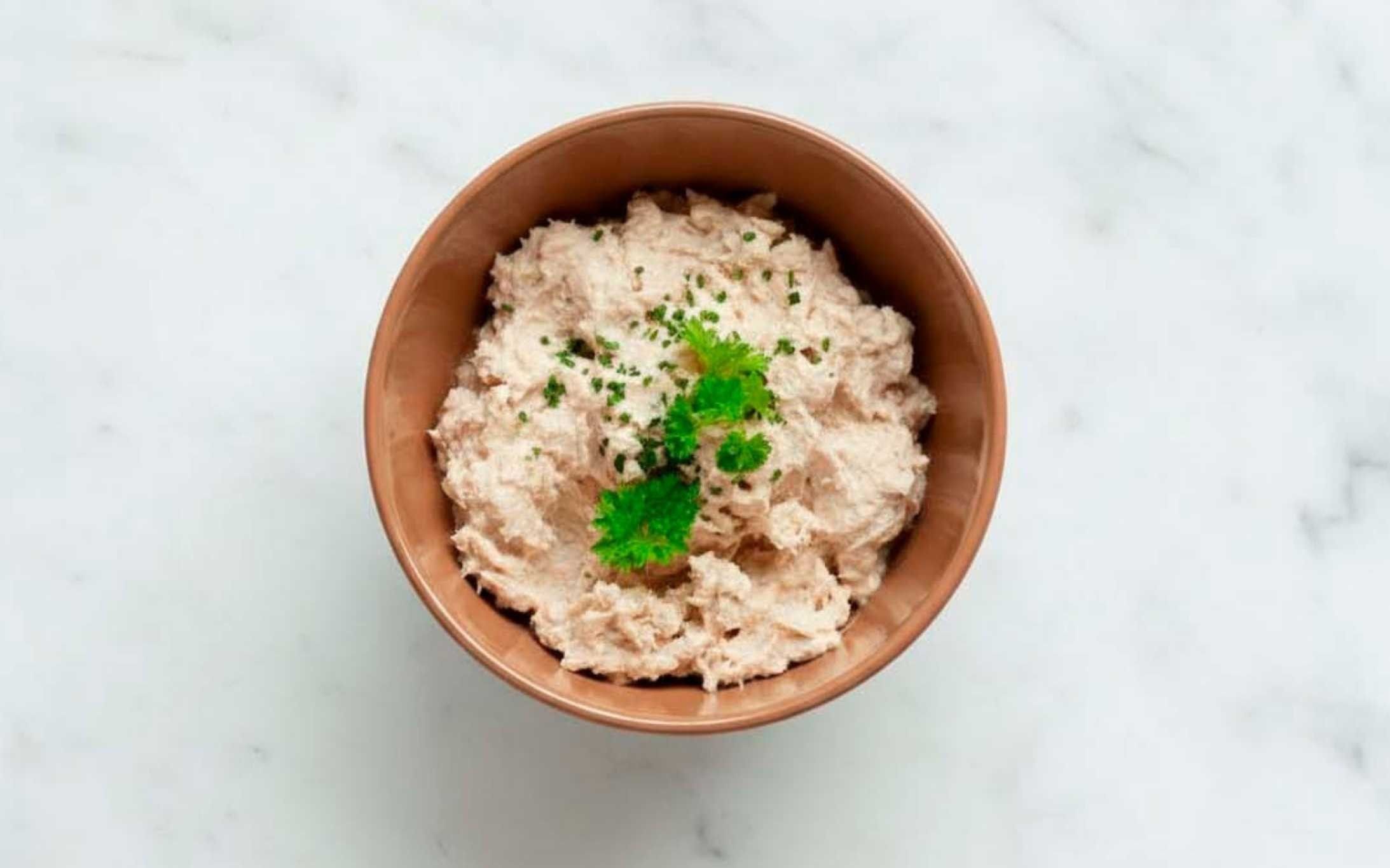 Tazón de cerámica con paté de atún cremoso decorado con cilantro fresco, sobre fondo de mármol blanco.