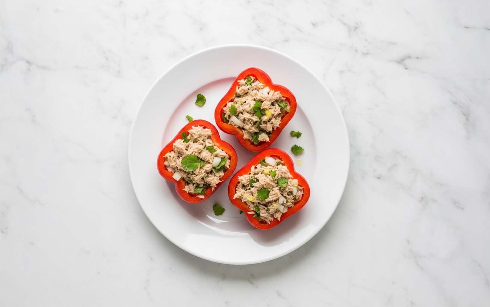 Tres mitades de pimiento rojo rellenas de atún con cebolla y cilantro, servidas en plato blanco sobre mármol.