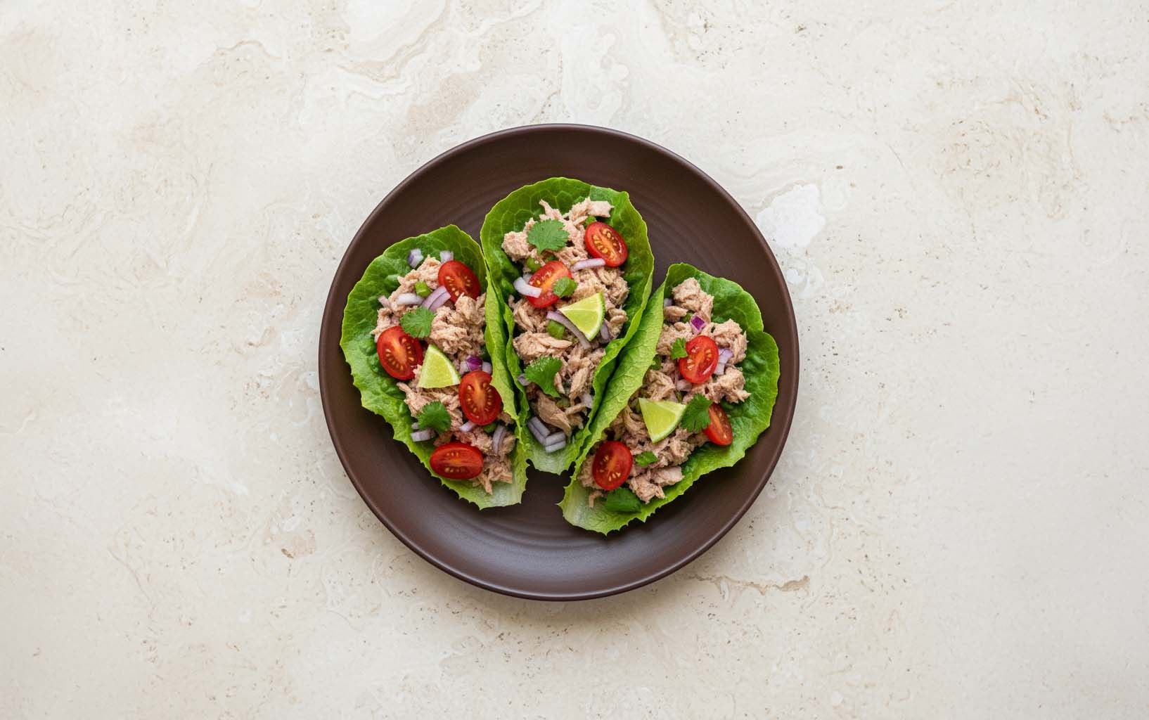 Tres tacos de lechuga rellenos de atún, tomate cherry, cebolla morada y cilantro con limón, en plato oscuro.