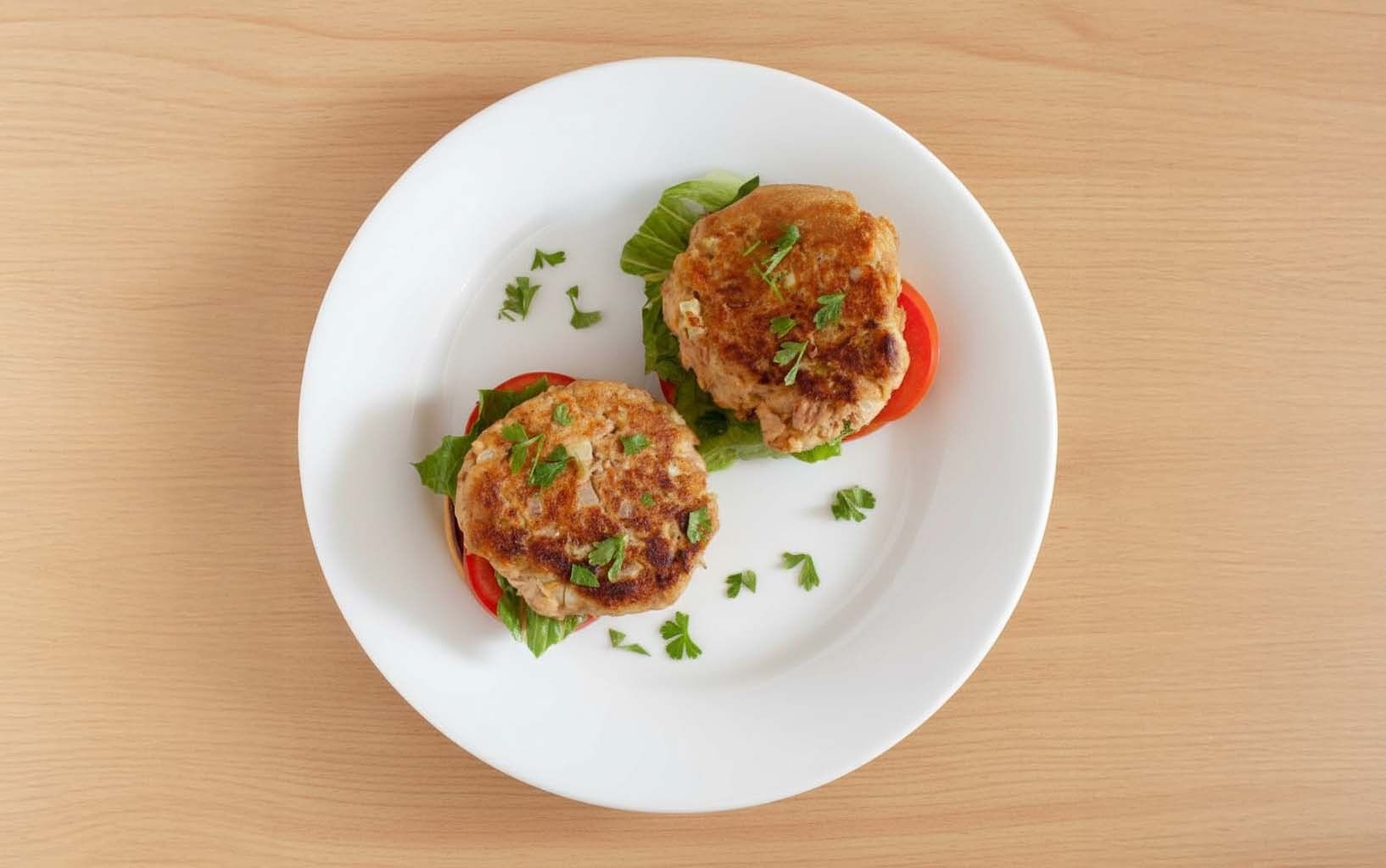 Dos torrejas de atún doradas sobre rodajas de tomate y lechuga, decoradas con perejil picado, servidas en plato blanco.