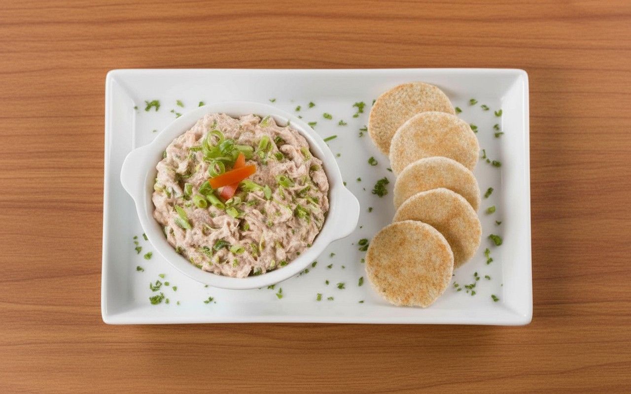 Dip cremoso de atún con cebolla china y pimiento en bowl blanco, acompañado de tortitas de avena redondas, servido en bandeja blanca sobre superficie de madera.