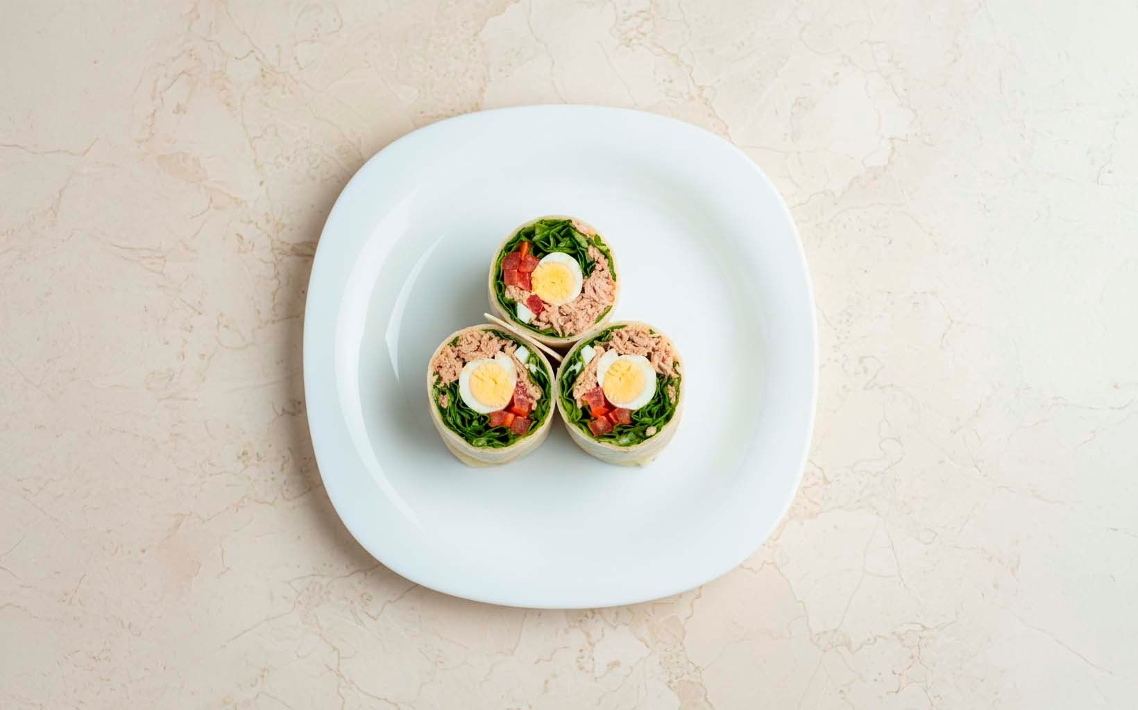Tres wraps de atún cortados al centro, rellenos de huevo duro, lechuga, tomate y atún, apilados en plato blanco.