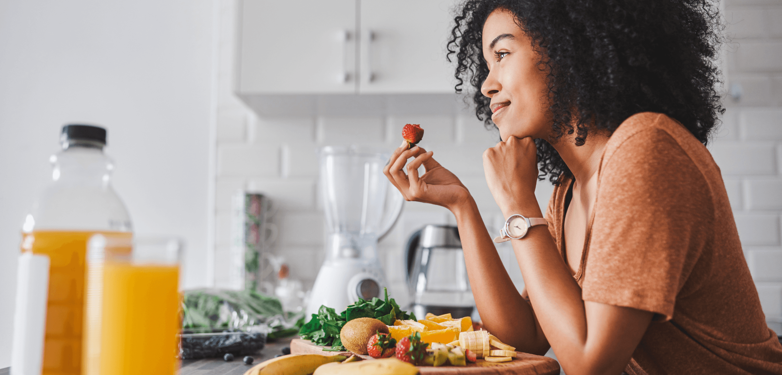 Woman smiling while eating a strawberry, surrounded by fresh fruits and vegetables in a bright kitchen, representing mindful eating and maintaining a healthy weight.